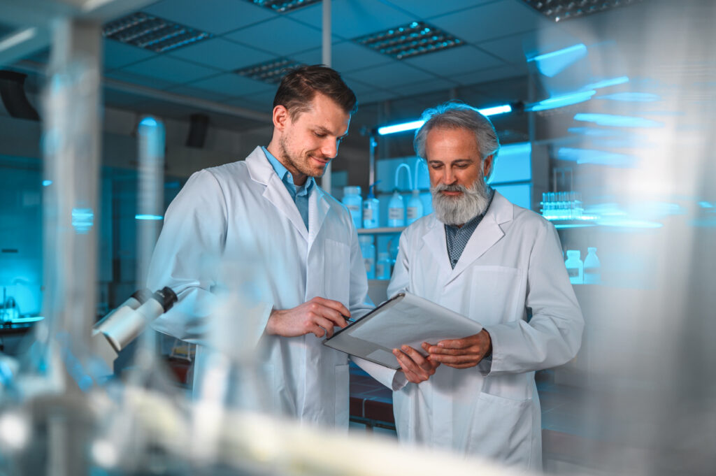 Male scientist with a beard and his younger intern standing in a laboratory, wearing white lab coats. Checking the results on a writing pad, smiling.  A  microscope visible. Candid waist up shot. Cold light. Blurred laboratory equipment in foreground.